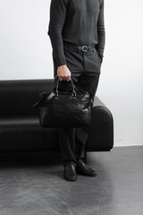 A professional man in a charcoal crewneck sweater and tailored trousers holds a black leather briefcase standing in a minimalist studio.