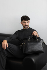 A professional male model in charcoal knitwear and tailored trousers sits on a black leather sofa holding a premium black leather briefcase.