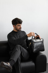 A professional male model in charcoal knitwear and tailored trousers sits on a black leather sofa holding a premium black leather briefcase.