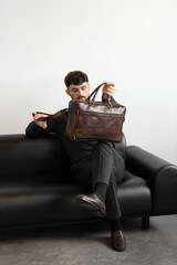 A professional male model in charcoal knitwear and tailored trousers sits on a black leather sofa holding a premium brown leather briefcase.