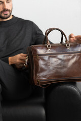 A professional male model in charcoal knitwear and tailored trousers sits on a black leather sofa holding a premium brown leather briefcase.