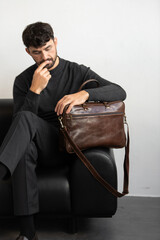 A professional male model in charcoal knitwear and tailored trousers sits on a black leather sofa holding a premium brown leather briefcase.