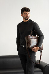 Professional male model in charcoal crewneck knitwear holding a dark brown leather briefcase in a minimalist studio setting