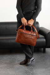 A professional man in a charcoal crewneck sweater and tailored trousers holds a light brown leather briefcase standing in a minimalist studio.