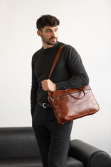 Professional male model in charcoal crewneck knitwear holding a light brown leather briefcase in a minimalist studio setting
