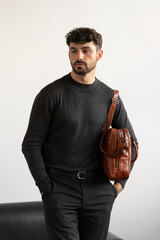 Professional male model in charcoal crewneck knitwear holding a light brown leather briefcase in a minimalist studio setting
