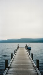 Fototapeta premium Long wooden pier extends into the calm lake water with a small blue boat gently moored at its end on a misty, tranquil day, creating a serene landscape.