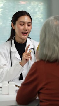 Young Asian physician in a white coat explaining the dosage of medication from a pill bottle to an elderly woman during a medical consultation in a modern hospital clinic room