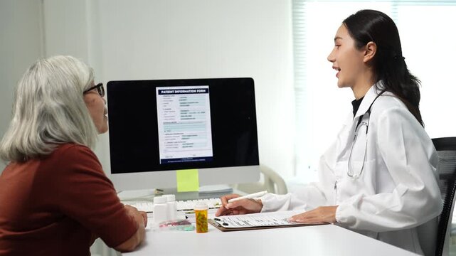 Female doctor in a white coat with a stethoscope talking to an elderly patient, pointing at a computer screen showing a patient information form and taking notes on a clipboard in her office