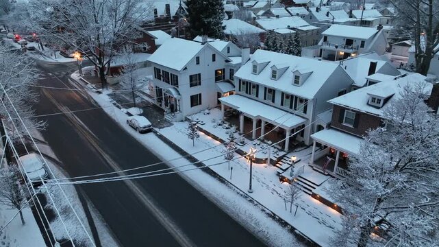 Aerial orbit shot of snow-covered American houses with decorated porches and warm lights, showing quiet suburban neighborhood during winter and Christmas season. Icy branches of trees.