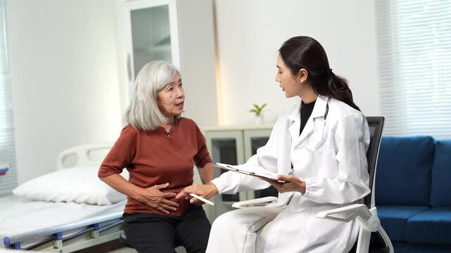 Elderly Asian woman talking with her doctor in the clinic, describing her stomach ache symptoms while the physician takes notes on a clipboard during a medical consultation and examination