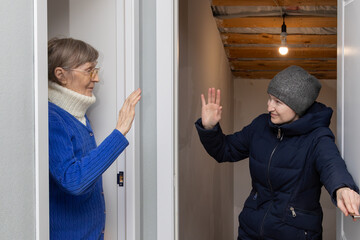 Woman dressed in winter clothes says goodbye to her elderly mother through an open door, waving goodbye before going outside.