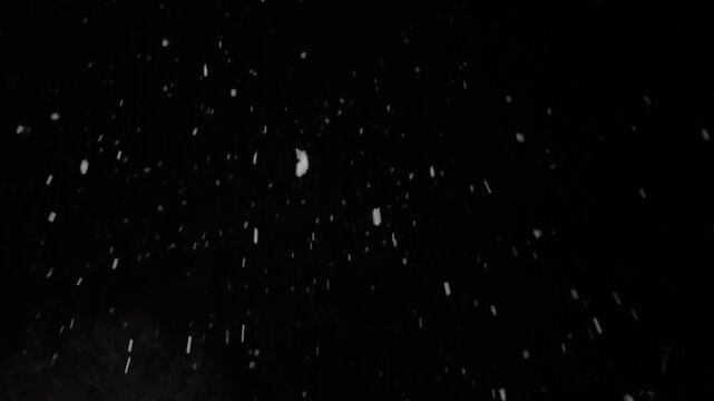 thick snow falls at night in the light of a streetlamp, snowflakes swirl in the wind and rush away against a black background.