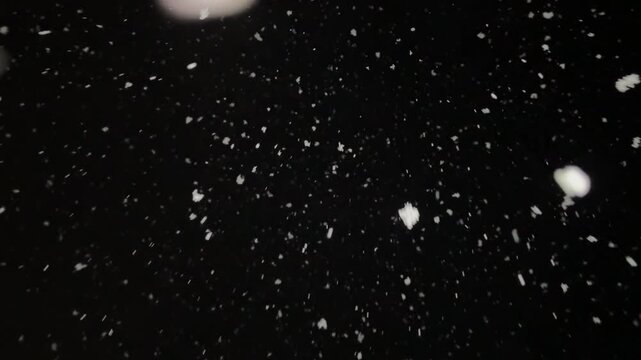 thick snow falls at night in the light of a streetlamp, snowflakes swirl in the wind and rush away against a black background.