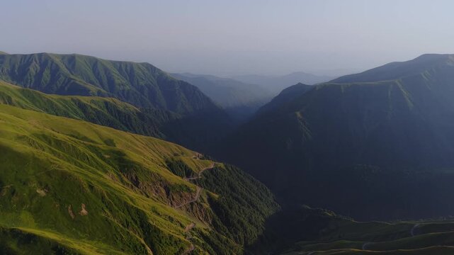 Aerial view of winding Alazani pass road through lush green mountain range, contrasting with a hazy sky, creating a scenic view, Chala, Tusheti, Georgia.