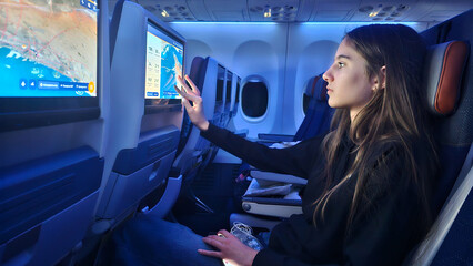 Teen Girl Using Inflight Entertainment Screen on Airplane. A young passenger interacts with a touchscreen seat monitor while flying in economy class during a night flight with blue lighting.