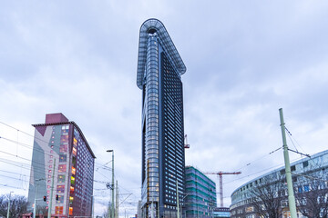 View of a towering skyscraper piercing the overcast sky, flanked by colorful buildings and a construction crane, creating a mix of modern and urban, The Hague, South Holland, Netherlands.