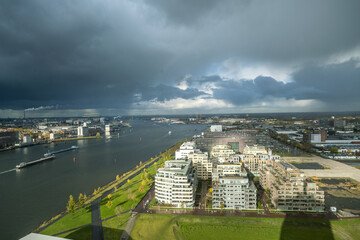 Aerial view of the IJ river reflecting the cloudy sky, lined with modern buildings and green spaces contrasting with the industrial landscape beyond, Amsterdam, North Holland, Netherlands.