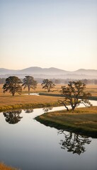 Naklejka premium Majestic trees and winding river through golden field at sunrise with misty mountains in the distance creating a peaceful natural landscape.