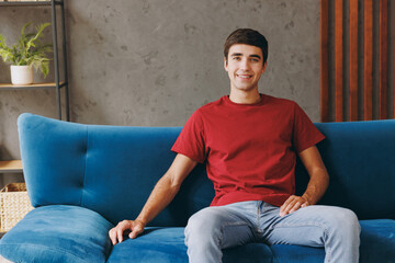 Young smiling cheerful happy fun european man wears red t-shirt sit on blue sofa couch stay at home hotel flat rest relax spend free spare time in living room indoors grey wall. People lounge concept.