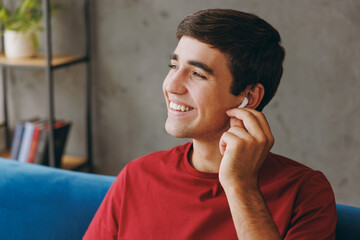 Young man wear red t-shirt listen music in earphones hold hand on gadget sit on blue sofa couch...