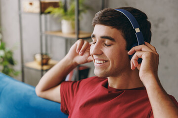 Close up calm young dreamful man wear red t-shirt headphones listen to music look aside sit on blue...