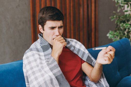 Young sick unhappy ill sad man 20s wears red t-shirt wrapped in plaid hold thermometer caugh covering mouth sit on blue sofa couch stay at home hotel flat spend time in living room indoor grey wall.