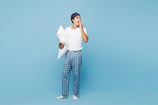 Full body calm young tired man he wearing pyjamas jam sleep eye mask rest relax at home yawn covering mouth with hand isolated on plain pastel light blue cyan background. Good mood night nap concept.