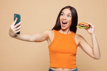 Fotobehang Muziek Young woman she wear orange casual clothes hold eat burger do selfie shot on mobile cell phone isolated on plain pastel beige background. Proper nutrition healthy fast food unhealthy choice concept.  © ViDi Studio
