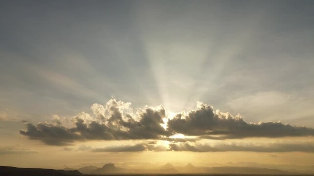 Aerial cinematic view of dramatic sunrise clouds with golden sun rays spreading across the sky above distant mountain silhouettes, creating a calm and atmospheric morning landscape.