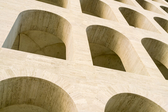 View of stark, geometric arches casting deep shadows on a textured travertine facade, a study in architectural repetition and sunlit stone, Rome, Lazio, Italy.