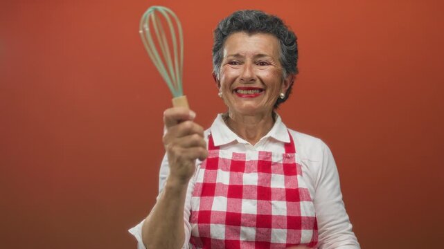 Senior woman with grey hair in a red checkered apron joyfully holds a whisk against an orange background, showcasing her passion for cooking and kitchen activities.