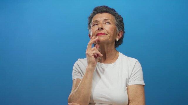 Senior woman with grey hair in white shirt thinking against blue background, expressing contemplation and curiosity with a thoughtful pose and a gentle smile.