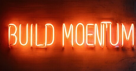 Bright neon sign displays text BUILD MOENTUM. Red glow contrasts dark textured wall. Simple, motivational message.