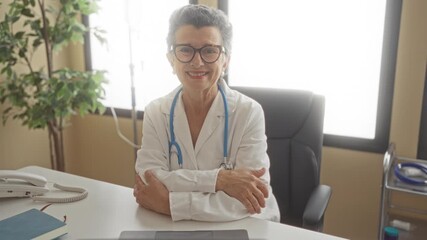 Senior female doctor with grey hair and glasses smiling confidently in a clinic office setting, with arms crossed, symbolizing professionalism and dedication to her medical practice.