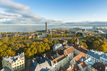 View of rooftops and trees ablaze with autumn colors meet the distant sea under a vast blue sky, a smokestack piercing the skyline, Tallinn, Harju County, Estonia.