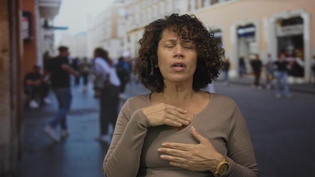 Woman with brown curly hair clutching chest on busy street amid blurred pedestrians by historic building facades; anxiety.
