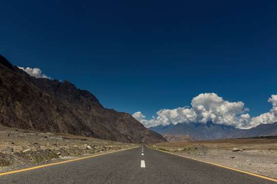 View of an endless asphalt ribbon slicing through the stark, rocky terrain beneath an azure sky dotted with fluffy clouds, Jaglot, Gilgit Baltistan, Pakistan.