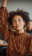 Portrait of confident young woman with curly hair and freckles wearing rust floral dress, hand raised, looking at camera with natural makeup and gold hoop earrings in bright interior