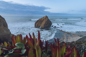 View of dramatic cliffs meet the foamy ocean, a massive rock formation stands against the horizon under a clouded sky, Santa Cruz, Lisboa, Portugal.