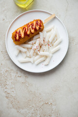 White plate with korean tteokbokki or garaetteok and corn-dog on a light-beige stone background, vertical shot with space, flat lay