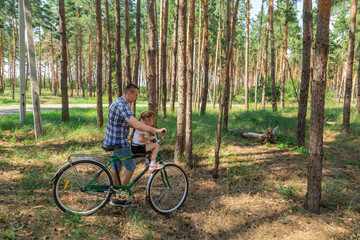 Fototapeta premium Dad rides his daughter in a bicycle seat in forest. A man carries a small child on a bicycle.