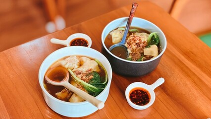 Two Bowls of Delicious Asian Wonton Noodle Soup with Fresh Greens and Chili Sauce on a Wooden Table