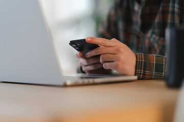 A person's hands, wearing a plaid shirt, hold a mobile phone, actively engaging with its screen. An open laptop is positioned on a wooden table in this indoor setting.