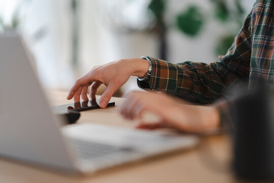 A person in a plaid shirt works at a desk, interacting with a laptop and a smartphone. They are focused on digital tasks, managing multiple devices simultaneously. This shows modern productivity.