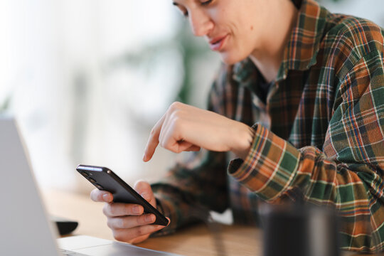 A young person in a green and brown plaid shirt is focused on their smartphone, tapping the screen with a finger. A laptop sits nearby on a wooden desk, suggesting a casual work or study environment.