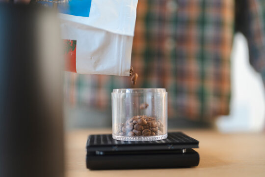 A person carefully pours fresh coffee beans from a white bag into a clear glass container resting on a digital scale. This precise measurement is essential for brewing quality coffee.