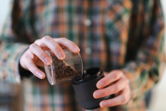 A person in a plaid shirt pours roasted coffee beans from a glass container into a manual coffee grinder. This is part of the morning coffee ritual.