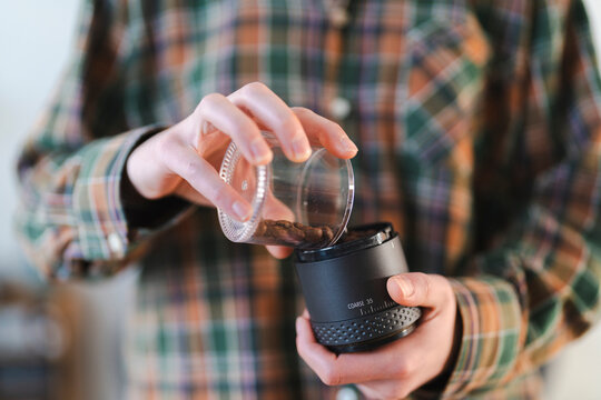 A person in a plaid shirt carefully pours whole roasted coffee beans from a clear glass container into a black manual coffee grinder. They are preparing to make fresh coffee.