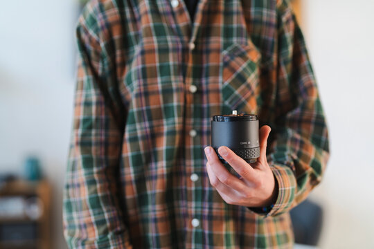 A person in a plaid shirt holds a manual coffee grinder. It is filled with whole coffee beans, ready for grinding. The grinder shows a 'COARSE 35' setting.